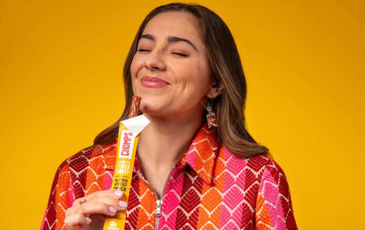 Woman smiling with a Chomps turkey stick against a yellow backdrop